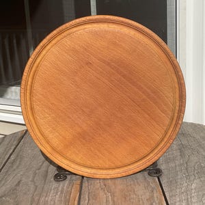 May include: A round wooden chopping board with a natural light brown grain and a smooth finish. The board features a circular carved groove running along the inner perimeter, providing a defined edge for the flat centre surface of the wood.
