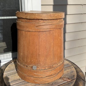 May include: A tall, cylindrical wooden bucket with a lid. The bucket is made of vertical wooden staves held together by several bands. The wood has a warm, reddish-brown tone. The bucket is sitting on a wooden spool.