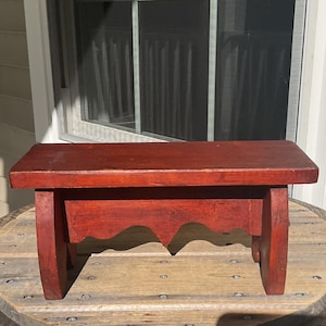 May include: A small, rectangular wooden stool with a deep reddish-brown finish. The stool features a flat top surface supported by two side panels, each designed with a decorative scalloped edge along the bottom, providing a stable base.