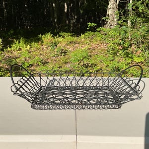 May include: A rectangular black wire basket featuring a decorative looped metal pattern along the sides and base. The basket includes two arched handles on opposite ends, constructed from thin, dark metal wire with a consistent open-weave design.
