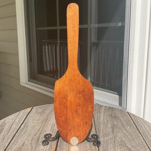 May include: A long, flat, wooden kitchen paddle with a rounded, spade-like base and a slender handle. The wood features a warm, medium-brown tone with visible natural grain patterns and a smooth, worn finish throughout the entire surface.
