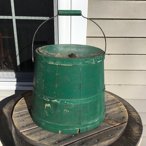 May include: A green wooden bucket with a metal handle and a wooden lid. The bucket is sitting on a wooden surface.