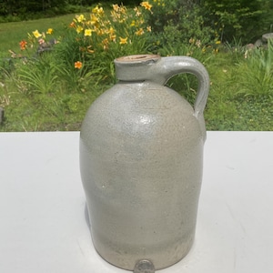 May include: A light gray stoneware jug with a single side handle and a narrow neck opening. The jug features a smooth, glazed finish and is positioned next to a standard U.S. quarter coin at the base for a clear sense of scale and proportion.