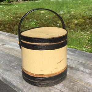 May include: A vintage wooden bucket with a black painted rim and handle. The bucket is painted a pale yellow colour and has a black painted band around the top and bottom.
