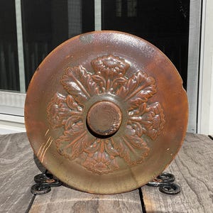 May include: A round, brown ceramic lid with a floral design in the center. The lid has a raised, circular handle and is resting on a decorative metal stand. The lid's color is a mix of brown and green.