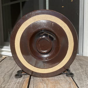 May include: A circular brown ceramic lid with a glossy finish and a wide, unglazed beige band around the center. The lid features a raised central knob and a small embossed number one near the bottom edge, showcasing a simple, functional design.