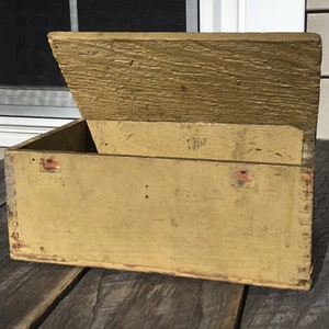 May include: A vintage wooden box with a hinged lid. The box is painted a pale yellow color and has a worn, rustic appearance. The lid is open, revealing the empty interior of the box.