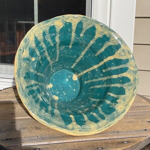 May include: A handmade ceramic bowl with a teal interior and a yellow rim. The bowl features a unique design of yellow streaks radiating from the center. The exterior is a light yellow color. This decorative bowl is perfect for serving or display.