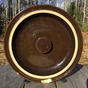 以下が含まれることがあります： 白い縁取りのある茶色の釉薬をかけた陶器の壺の蓋の裏側。蓋の中央には円形のくぼみがある。