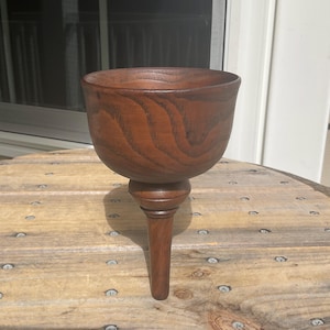 May include: A dark brown wooden goblet with a deep, rounded bowl and a prominent, tapered stem. The wood features a visible, swirling grain pattern throughout the surface, showcasing the natural texture and rich, warm tones of the material.