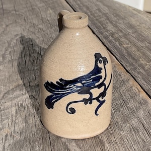 May include: A small, tan stoneware jug with a handle and a dark blue bird design. The bird is perched on a branch, and the jug has a rounded body and a narrow neck. The jug is likely vintage or antique.