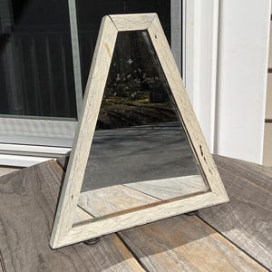 May include: A triangular mirror with a distressed, off-white wooden frame. The mirror is supported by small, round feet. The frame has a textured appearance, and the mirror reflects the outdoor surroundings.