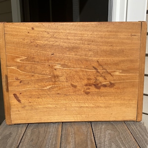 May include: A rectangular wooden box with a natural light brown finish, featuring visible wood grain patterns and various surface scratches, scuffs, and dark marks across the front panel, constructed with vertical side supports.