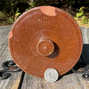 May include: A round, brown ceramic lid with a central knob. The lid has a slightly textured surface and is placed on a decorative metal stand. A quarter coin is next to the lid for size comparison. The lid has some areas where the brown paint has chipped off.