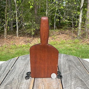 May include: A wooden butter paddle with a handle and a flat, rectangular blade. The paddle is a rich, reddish-brown colour and sits on a decorative metal stand. A coin is next to the paddle for size comparison.