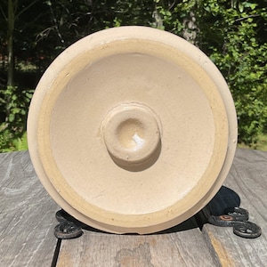 May include: A circular, light beige ceramic lid with a smooth, matte finish. The underside features a raised, centered circular knob or handle, and a wide, flat outer rim designed to fit securely onto a corresponding container or vessel.