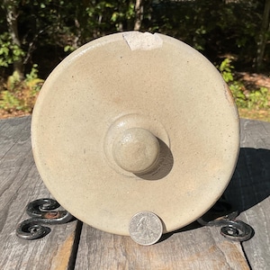 Peut inclure: Couvercle de poterie en grès beige avec un bouton arrondi, montrant des signes d'âge et d'usure. Le couvercle est posé sur un support décoratif en fer forgé. Une pièce de monnaie américaine est à côté du couvercle pour la comparaison de taille.