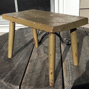 May include: A rustic wooden stool with four legs, painted in a light green color. The stool is weathered and shows signs of age. It is sitting on a wooden surface.