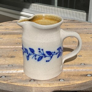 May include: A stoneware pitcher with a speckled off-white glaze and a brown interior. The exterior features a raised blue relief pattern of leaves and berries encircling the middle. It has a curved handle and a wide pouring spout.