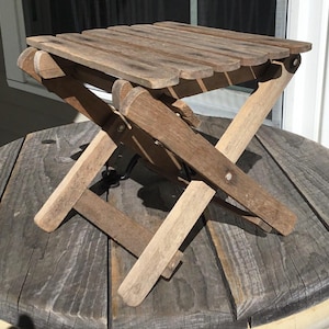 May include: A small, folding wooden side table with a slatted top. The table is made of weathered wood and has an X-shaped leg design. The table is approximately 12 inches square and 12 inches high.