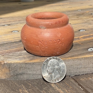 May include: A small, round, terracotta-coloured ceramic pot with a wide opening and a thick rim. The side of the pot features the embossed text Necco Boston Baked Beans. A silver-coloured United States quarter coin is placed in front for scale.