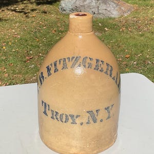May include: A tan stoneware jug with a narrow neck and a cylindrical body. The front features dark blue stenciled text that reads J.B. FITZGERALD and TROY, N.Y. The surface has a smooth, glazed finish with a visible rim at the top opening.