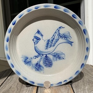 May include: A round, off-white stoneware pie dish featuring a hand-painted blue cardinal bird perched on a pine branch with a pinecone. The rim is decorated with a repeating pattern of blue teardrop shapes. A US quarter coin is placed at the bottom.