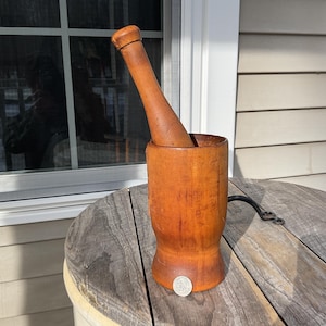 May include: A wooden mortar and pestle set. The mortar is a cylindrical, orange-brown colour, with a matching pestle. The set is displayed on a weathered wooden table. A silver coin is at the base of the mortar.