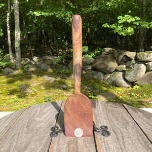 May include: A brown wooden spatula with a long, rounded handle and a wide, flat blade. The spatula is held upright by a black metal stand with decorative scrollwork at the base. A silver coin is placed at the bottom centre of the wooden blade.