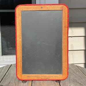 May include: A vintage chalkboard with a wooden frame and red trim. The chalkboard has a dark grey surface and the frame is made of light brown wood. The text "FROM THE ORIGINAL VERMONT COUNTRY STORE" is printed at the bottom.