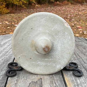 May include: A light gray stoneware crock lid with a central knob, resting on a decorative black metal stand. The lid has a circular shape with a slightly rough texture and visible imperfections. The stand has a scrolled design.