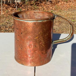 May include: A copper mug with a large handle. The mug has a cylindrical shape and a slightly flared rim. The copper has a warm, reddish-brown color with areas of green oxidation. The handle is curved and attached to the side of the mug.