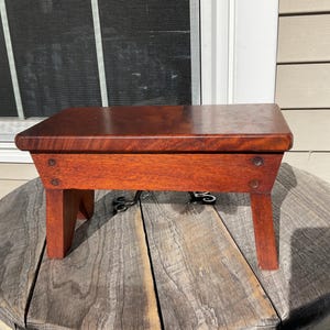 May include: A small, wooden stool with a dark reddish-brown finish. The stool has a rectangular top and four legs. The legs are slightly angled inward.