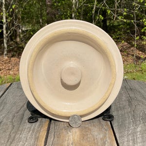 May include: A beige stoneware crock with a circular design and a central knob. The crock is set on a black metal stand. A coin is placed near the base for size reference. The crock has a rustic, vintage aesthetic.