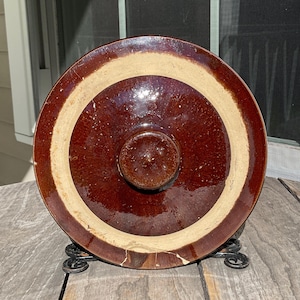 以下が含まれることがあります： ベージュと茶色の同心円模様の茶色の陶器の蓋。 蓋には中央のノブがあり、装飾的な金属製のスタンドに表示されています。 蓋は光沢仕上げで、おそらくヴィンテージです。