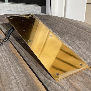 May include: A gold-colored, triangular door knocker with a decorative, wrought-iron handle. The door knocker is made of a shiny, reflective metal and is mounted on a wooden surface. The handle has a scrolled design.