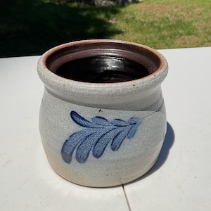 May include: A round stoneware crock featuring a speckled light gray exterior with a dark brown interior rim. The front of the vessel is decorated with a single blue hand-painted fern leaf motif. The pot has a wide opening and a thick, rounded lip.
