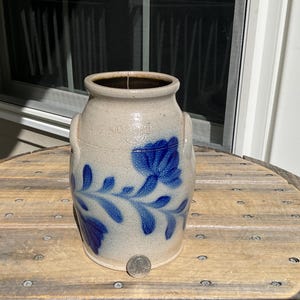 May include: A beige stoneware crock featuring a blue hand-painted floral design with leaves and a central flower. The vessel has two side handles and a wide opening. A silver-colored coin is placed at the base for scale, showing the size.