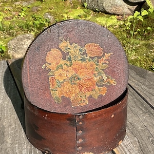 May include: A circular antique wooden box with a lid. The lid displays a floral design in orange, yellow, and green hues on a dark brown backdrop. The box is a dark brown colour and seems to be constructed from wood.