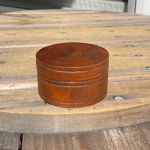 May include: A small, round, lidded container crafted from polished brown wood. The object features a smooth, flat circular top and a cylindrical base with a visible horizontal seam where the lid meets the body, showcasing a natural wood grain.