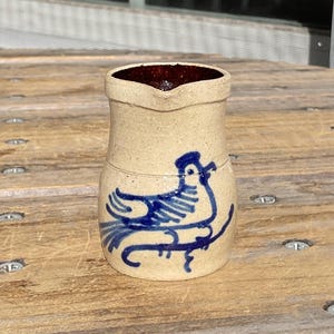 May include: A small, cream-colored ceramic pitcher with a blue bird design. The pitcher has a brown rim and a slightly flared lip.