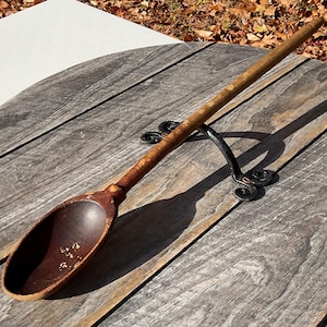 May include: A long wooden spoon with a deep, oval bowl and a slender handle. The spoon is a dark brown colour with visible wear and tear, suggesting age and use. It rests on a decorative black metal stand, set against a weathered wooden surface.
