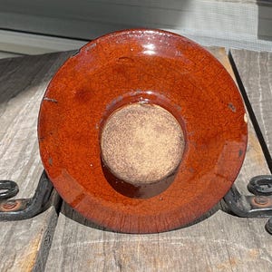 May include: A round, reddish-brown ceramic lid with a central knob, resting on a decorative wrought iron stand. The lid has a cracked glaze effect. The stand features scrolled feet.