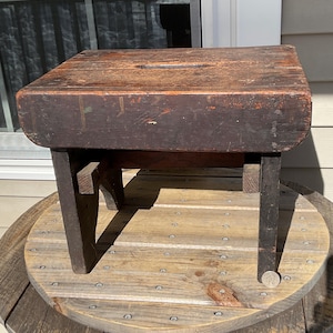 May include: A small, antique wooden stool with a rectangular seat and four sturdy legs. The stool is stained a dark brown color, showing signs of wear and age. A small, rectangular cutout is in the center of the seat. The stool is approximately 12 inches tall.