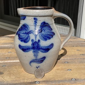 May include: A speckled light gray stoneware pitcher featuring a cobalt blue hand-painted floral design on the front. The pitcher has a curved handle and a wide spout. A silver-colored United States quarter coin is placed at the base for scale.