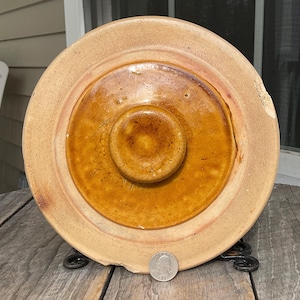 以下が含まれることがあります： 中央にノブが付いた丸いアンティークの炻器の蓋。蓋には、茶色とタンの色合いの同心円があり、端がわずかに欠けています。サイズの目安として、蓋の近くにコインが置かれています。