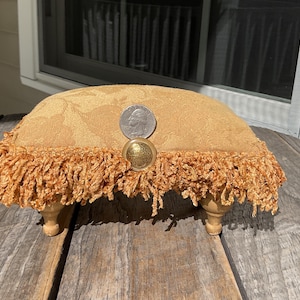 May include: A small, upholstered footstool with a gold floral pattern and fringe trim. The stool has four wooden legs. A quarter and a decorative button are on top of the stool.