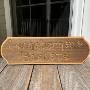 May include: A rectangular wooden sign with rounded, decorative edges featuring the carved text Longaberger Baskets and a registered trademark symbol. The sign has a dark brown stained finish with a lighter natural wood border around the perimeter.