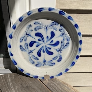 May include: A round, light grey ceramic pie dish featuring a hand-painted cobalt blue dragonfly motif in the centre, surrounded by a swirling petal pattern. The rim is decorated with a repeating pattern of small, blue, teardrop-shaped marks.