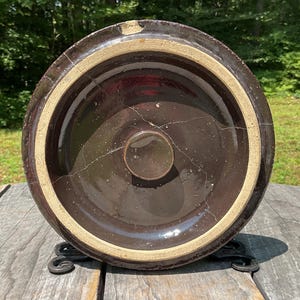 以下が含まれることがあります： ベージュの縁と小さな円形の取っ手が付いた、濃い茶色のセラミック製のクロック蓋。蓋にはひび割れが見え、古くなっているように見えます。クロック蓋は、キッチンや家の装飾用である可能性があります。